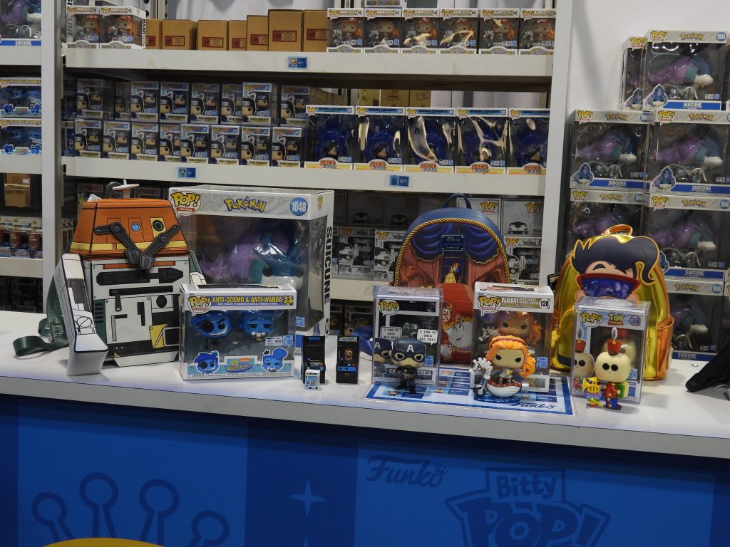 a booth with several Funko pops in the background. On the table are the Chopper Loungefly backpack, Beauty and the Beast ballroom scene backpack, Goofy backpack, the Suicune Funko pop, Anti-Cosmo and Anti-Wanda two pack Funkos, Pixar's Tin Toy Funko, Captain America from the Infinity Saga Funko, One Piece's Nami with a white hobby horse, and two of the Bitty Arcade Pops (Tron and Wreck-It Ralph).