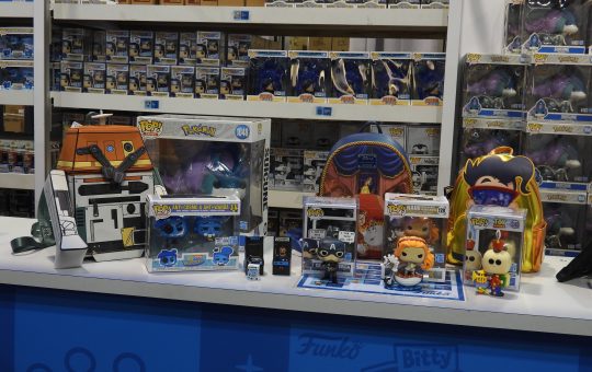 a booth with several Funko pops in the background. On the table are the Chopper Loungefly backpack, Beauty and the Beast ballroom scene backpack, Goofy backpack, the Suicune Funko pop, Anti-Cosmo and Anti-Wanda two pack Funkos, Pixar's Tin Toy Funko, Captain America from the Infinity Saga Funko, One Piece's Nami with a white hobby horse, and two of the Bitty Arcade Pops (Tron and Wreck-It Ralph).
