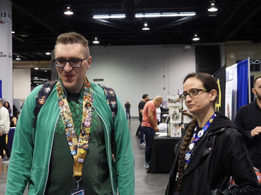 a white man with short brown hair with glasses, wearing a green hoodie over a black t-shirt with a green design and a lanyard with a lot of pins, standing next to a nonbinary white person with brownish-red hair with a braid and piercings in the ear and on the bottom lip, wearing a black pleather jacket