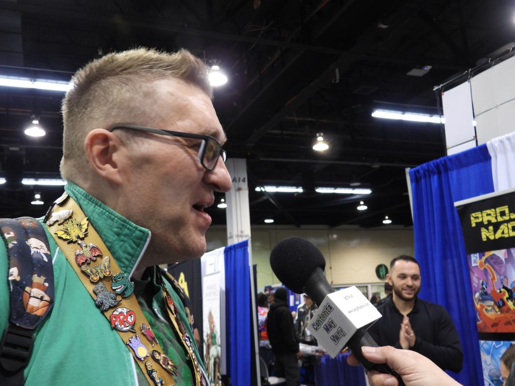 a white man with short brown hair with glasses, wearing a green hoodie over a black t-shirt with a green design and a lanyard with a lot of pins talking into a microphone