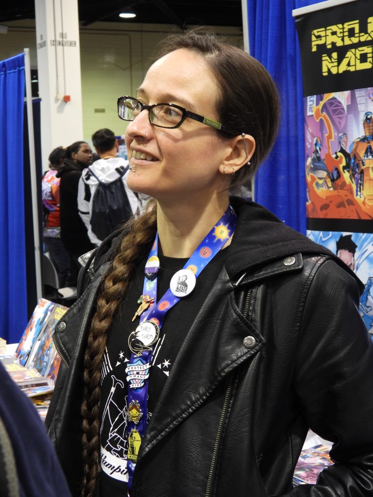 a nonbinary white person with brownish-red hair with a braid and piercings in the ear and on the bottom lip, wearing a black pleather jacket