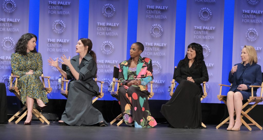 Five women sit in director-style chairs. From left to right: a white woman with dark hair, wearing a dress that is green and yellow; a white woman with long brown hair in a pigtail, wearing a gray trench coat and gesturing; a black woman with buzzed black hair, wearing an outfit that's black with batches of other colors; an Asian woman with shoulder-length black hair, wearing a black dress; and a white woman with blond hair; wearing a blue skirt-suit.