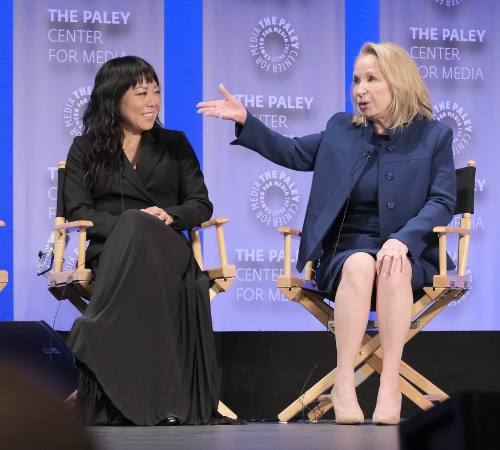 Two women sitting on director-style chairs. An Asian woman with shoulder-length black hair, wearing a black dress and a white woman with blond hair; wearing a blue skirt-suit.