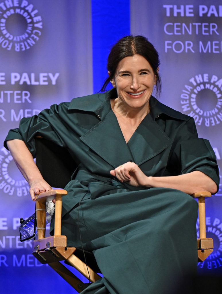 a white woman with long brown hair in a pigtail, wearing a gray trench coat, sitting in a director-style chair
