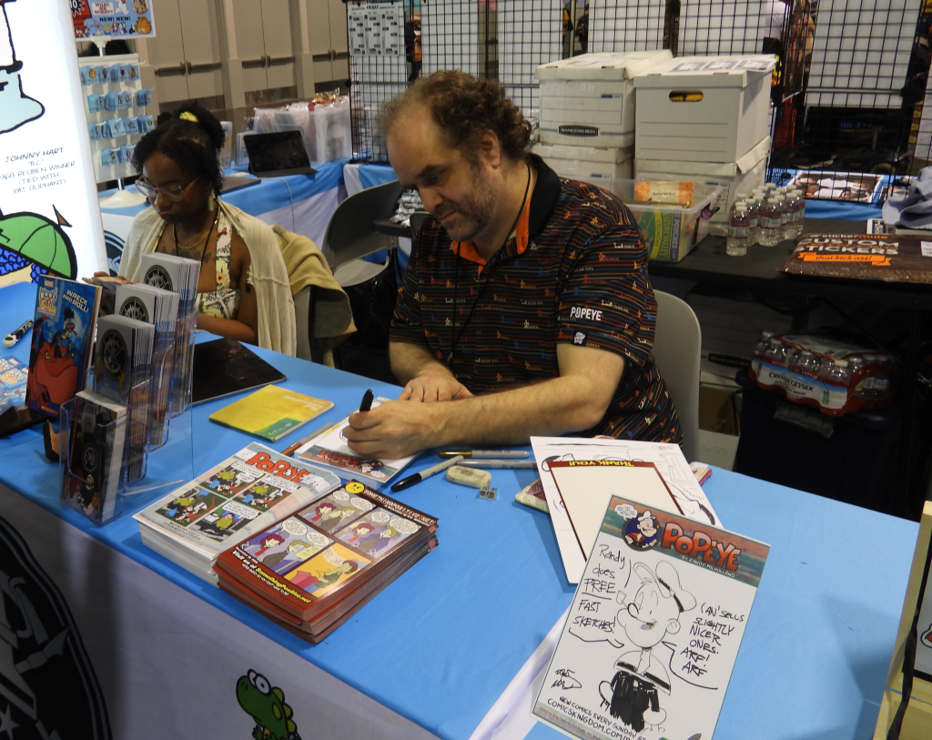 a young black woman sitting at a table next to a middle aged white man with brown wearing a Popeye shirt and drawing
