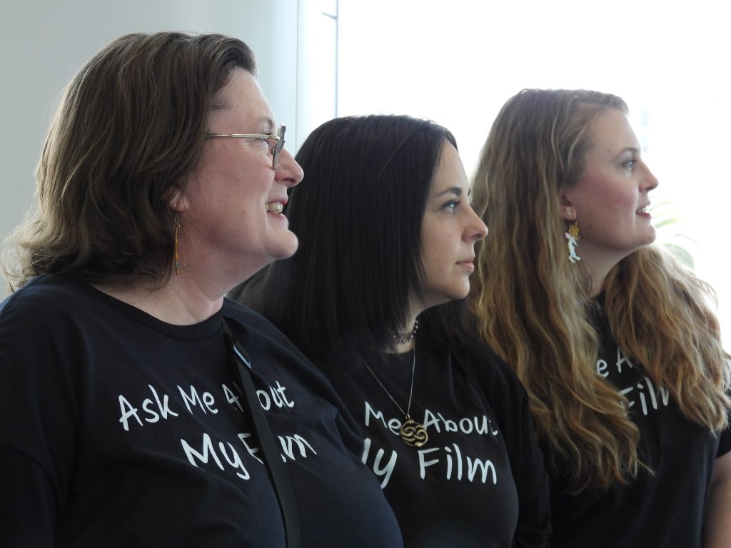 Three white women, one in middle aged and the other two in their twenties, smiling and looking off camera. They all wear shirts that say 'Ask Me About My Film'