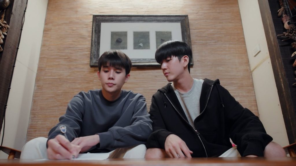 Thap and In sit side by side on a couch. Thap is writing on the list of boundaries on the coffee table in front of him while In looks over his shoulder at what he's writing.