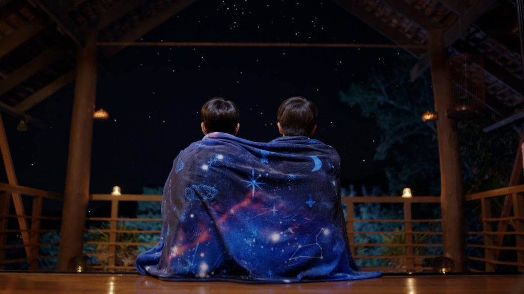 A behind shot of In and Thap, sitting closely side by side, wrapped under In's space blanket. They sit on the porch, looking up at the night sky.