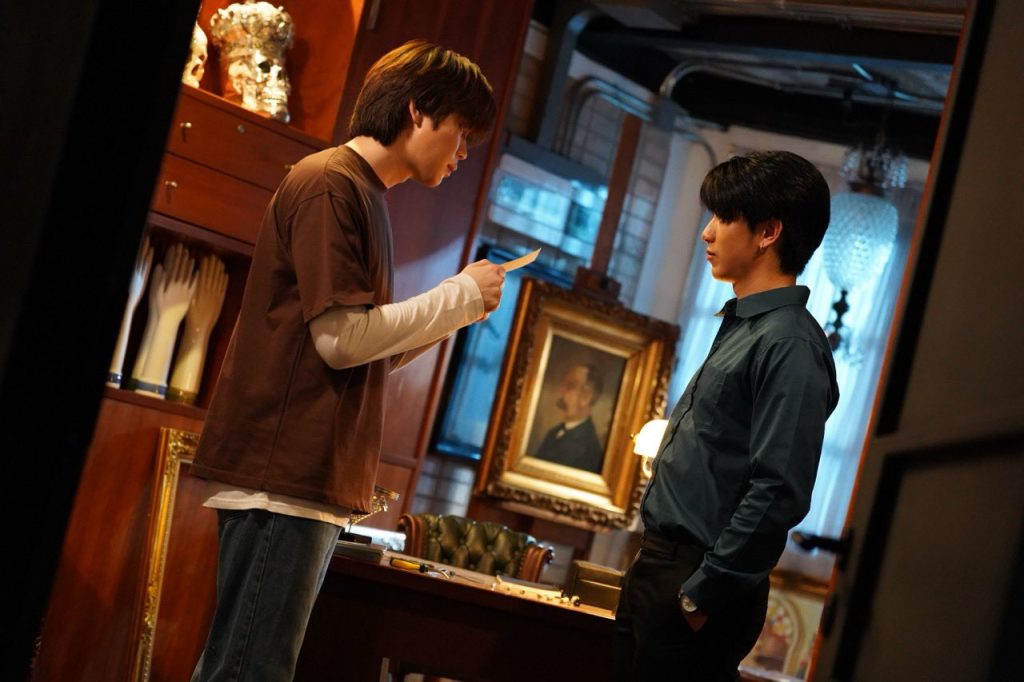 Punn and Jett stand opposite each other in Punn's antique shop. Jett has his hands in his pockets. Punn is holding a photo with both hands (you can't see it, but the photo is of the ruined painting of Ramil).