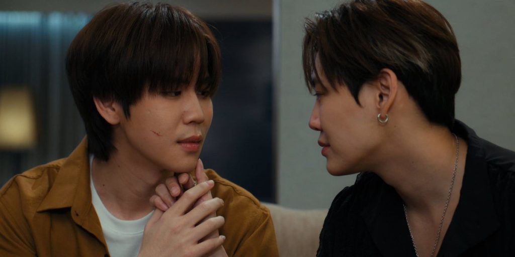 Punn and Ramil sit on the sofa, angled towards each other. Ramil has his hand on Punn's chin, examining a wound on Punn's check. Punn is holding Ramil's hand.