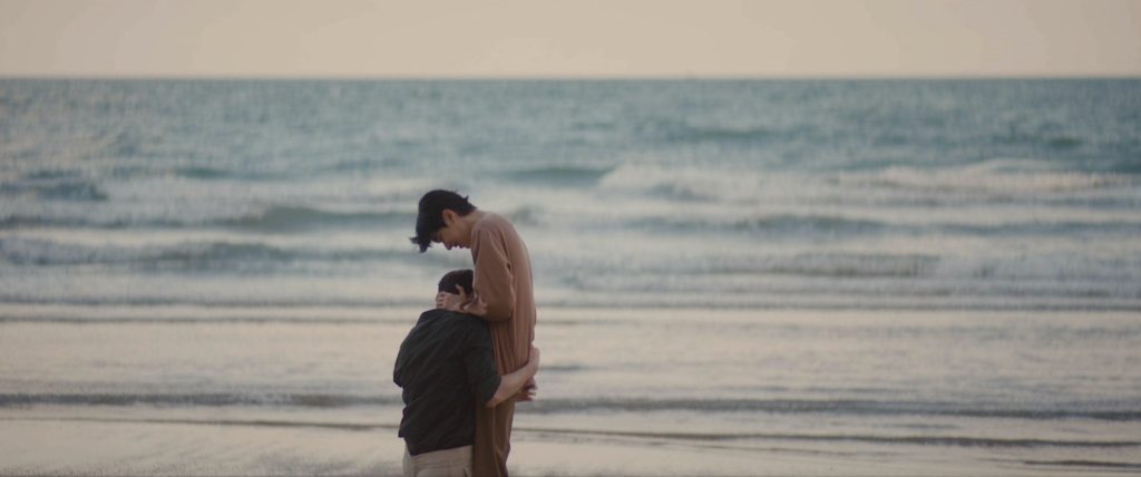 Ramil kneels in front of Paytai on the beach, his arms wrapped around him, and his head buried in his stomach.