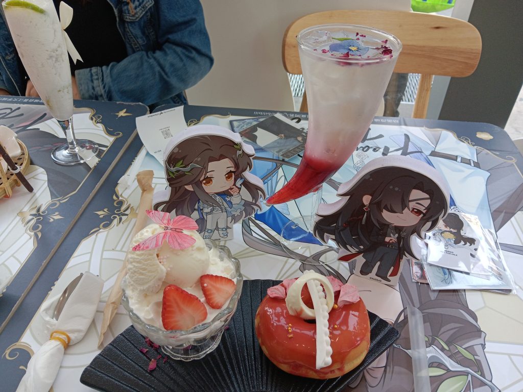 The "Token of Trust" is a donut with strawberry glaze and dish of vanilla ice cream served on a plate shaped like a folding fan. A butterfly sits on top of the ice cream. The donut is covered with a chocolate version of San Lang's ring. The "Elation" is a mixed berry soda served in a horn-shaped glass.