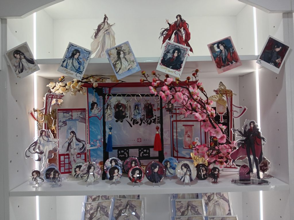 Merch display features pins, keychains, acrylic standees, and photo cards, as well as decorations like cherry blossoms.