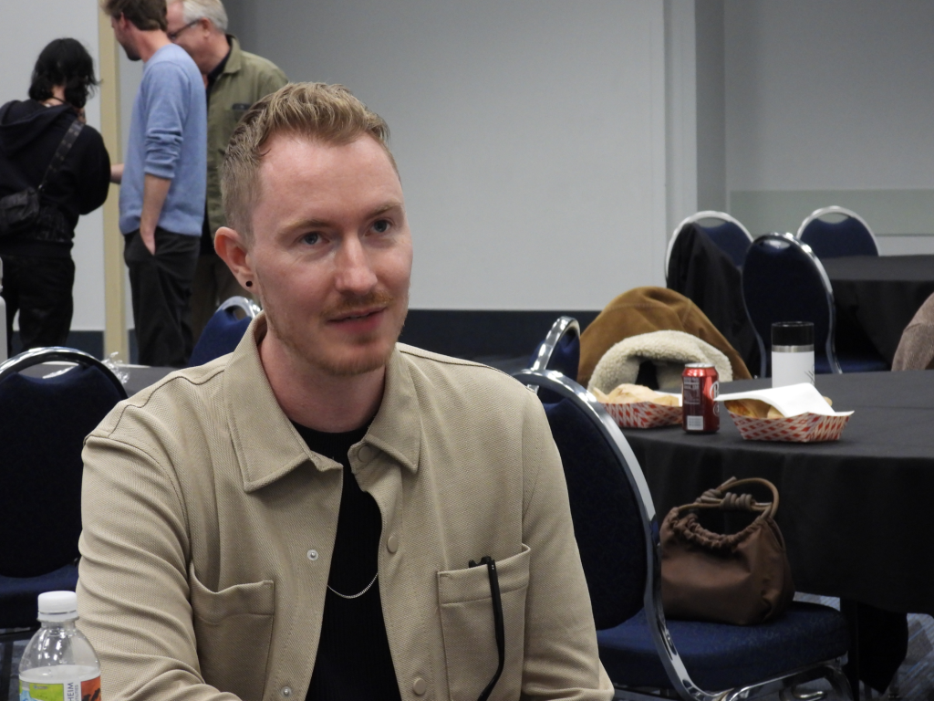 a white man with light brown/reddish hair and a slight mustache wearing a tan button up jacket over a black shirt