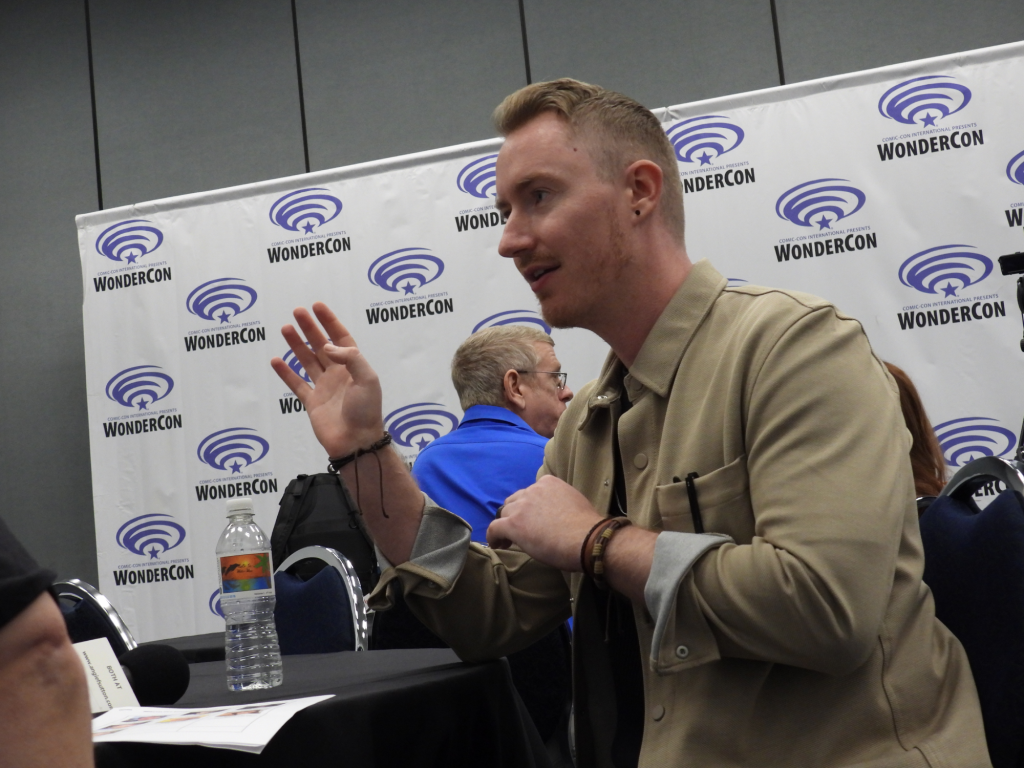 a white man with light brown/reddish hair and a slight mustache wearing a tan button up jacket over a black shirt