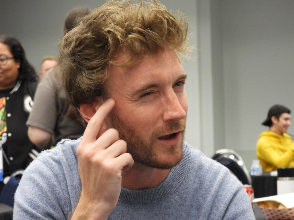a white man with reddish brown hair and beard, wearing a grey sweatshirt, squinting and pointing to his forehead