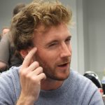 a white man with reddish brown hair and beard, wearing a grey sweatshirt, squinting and pointing to his forehead