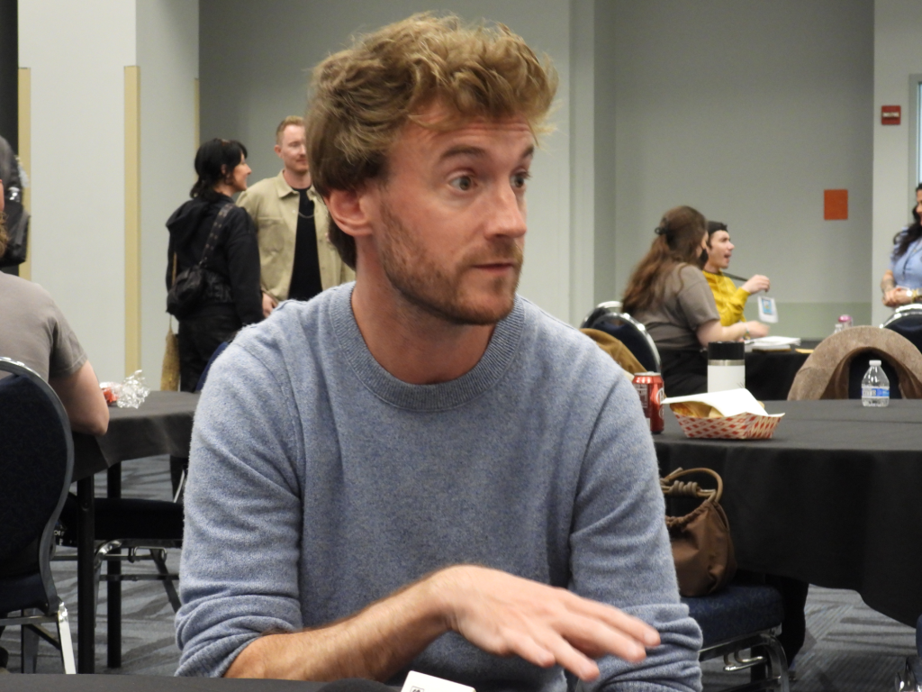 a white man with reddish brown hair and beard, wearing a grey sweatshirt, eyes wide open with one hand out
