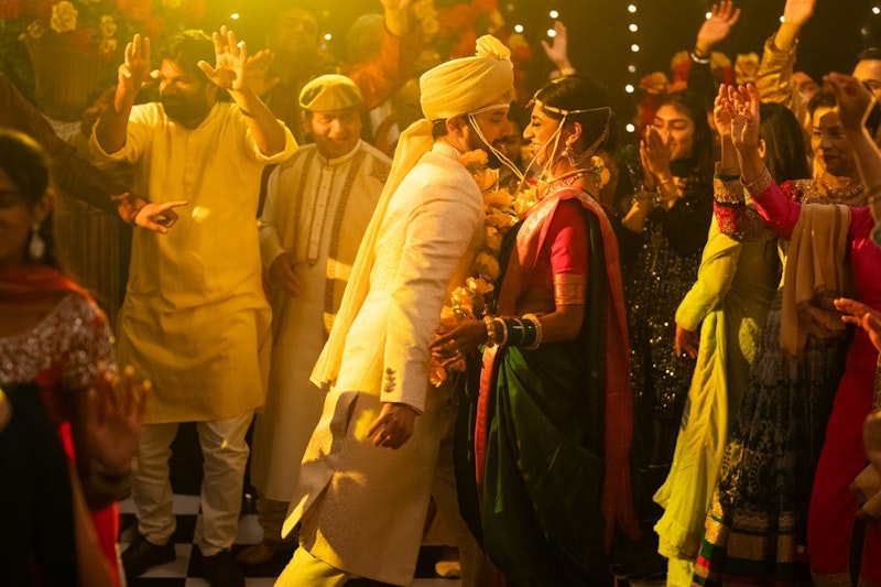 A scene of an Indian wedding. Many people circle a man and woman couple in dance. The man is in white with a turban, while the woman is in a red and green sari. 