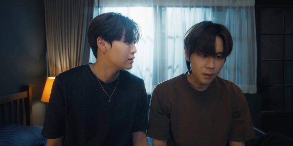 Ramil and Punn sit side by side on Punn's bed. Punn is staring off in the distance while Ramil is turned to look at him. Both look extremely upset.