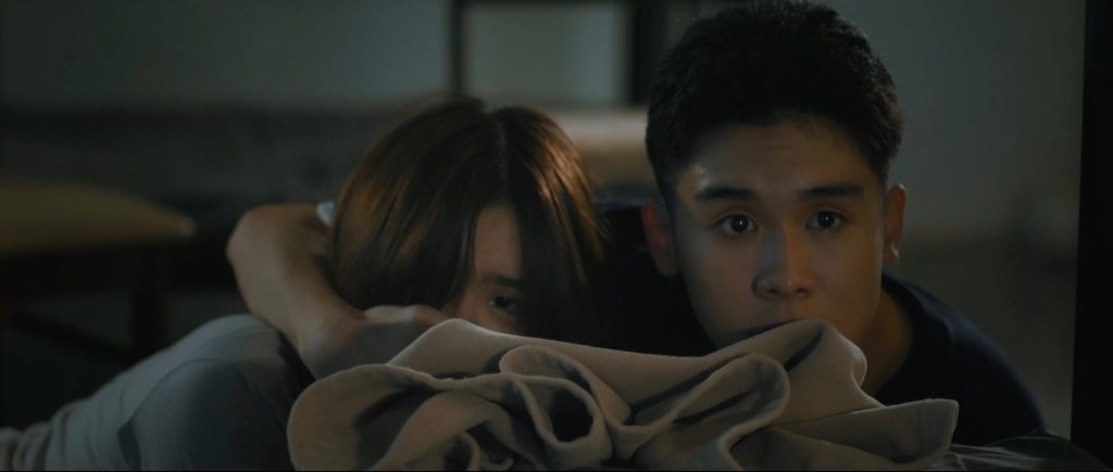 Teenage Punn and Elise hide behind a bed as their orphanage is attacked. Punn is covering Elise's mouth with his hand.