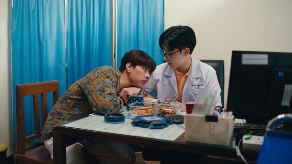 Peng and Wut are eating lunch at Wut's desk in his office. Peng has his arms wrapped around Wut's arm, leaning his head against him, staring up at him.