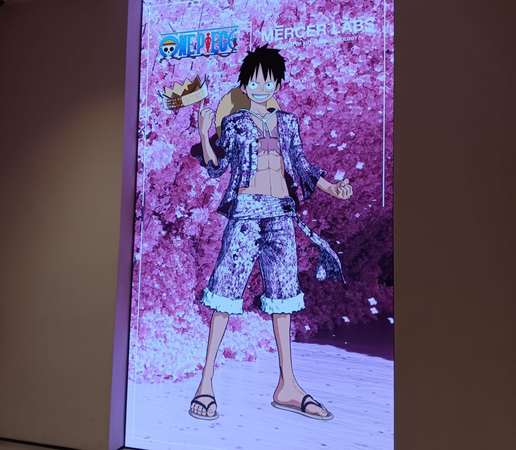 The One Piece immersive exhibit key art, which is at the entrance to the exhibit. Luffy stands proudly in a cave where the walls are made of sakura blossoms. His outfit matches the walls - pink and patterned like cherry blossoms. He is twirling a crown on his finger.