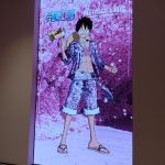 The One Piece immersive exhibit key art, which is at the entrance to the exhibit. Luffy stands proudly in a cave where the walls are made of sakura blossoms. His outfit matches the walls - pink and patterned like cherry blossoms. He is twirling a crown on his finger.