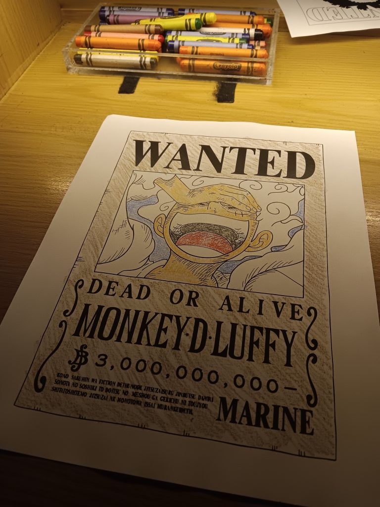 A photo of the Monkey D. Luffy wanted poster that Jamie has colored. The poster is on a table with the tray of crayons above it.