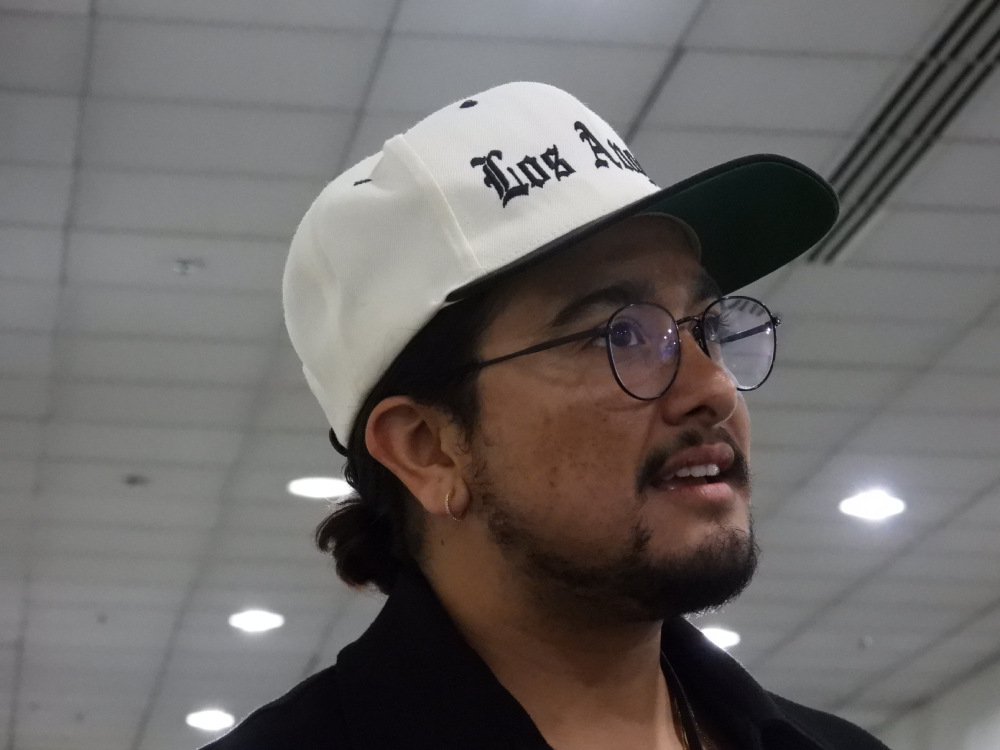 a Latiné transmasculine nonbinary person with dark hair and a slight beard, wearing a white Los Angeles hat, glasses, and a black top