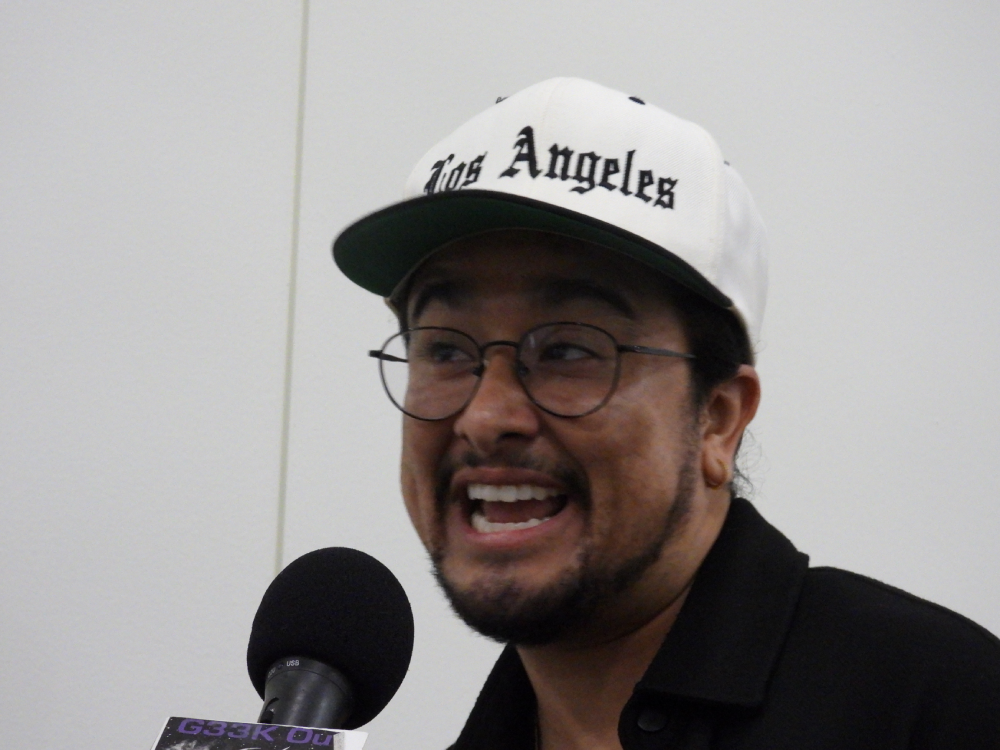 a Latiné transmasculine nonbinary person with dark hair and a slight beard, wearing a white Los Angeles hat, glasses, and a black top