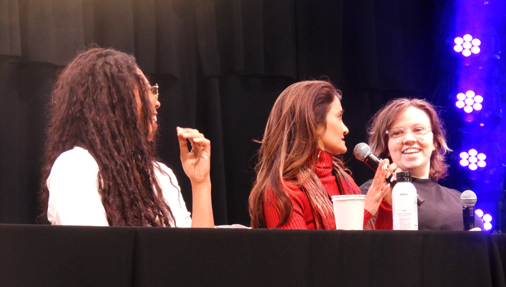 three people sitting at a table; a black woman with long black hair and glasses wearing a white top; a white woman with long brownish-blond hair wearing red turtleneck; and a white person with short brown hair, glasses, and a black top