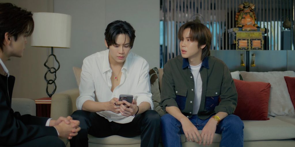 Punn and Ramil sit side by side on the sofa. Methas sits in an arm chair at an angle from Ramil. Punn and Methas are looking at each other while Ramil stares down at Methas's phone.