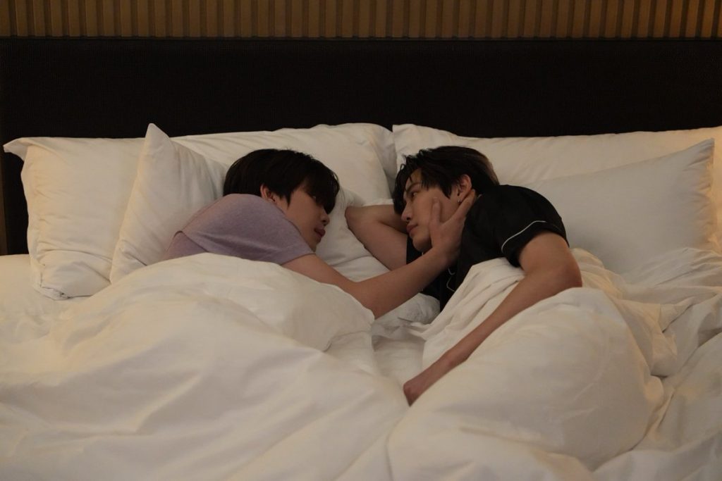 Punn and Ramil lie side by side facing each other in bed. Ramil has one arm bent underneath his head. Punn is resting his hand on Ramil's cheek.