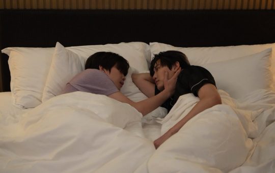 Punn and Ramil lie side by side facing each other in bed. Ramil has one arm bent underneath his head. Punn is resting his hand on Ramil's cheek.
