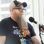 a white man with a long reddish curly beard starting to turn white, wearing glasses, a baseball cap, and a black t-shirt, being interviewed