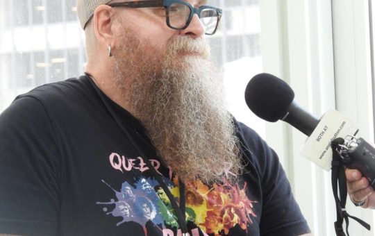a white man with a long reddish curly beard starting to turn white, wearing glasses, a baseball cap, and a black t-shirt, being interviewed
