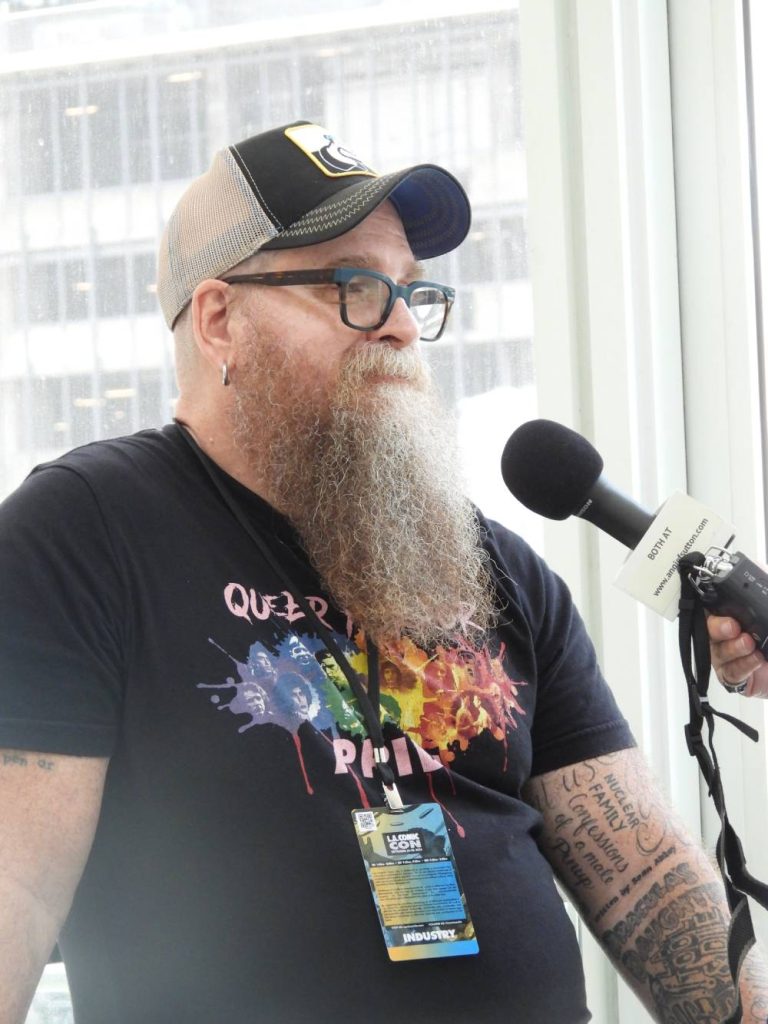 a white man with a long reddish curly beard starting to turn white, wearing glasses, a baseball cap, and a black t-shirt, being interviewed