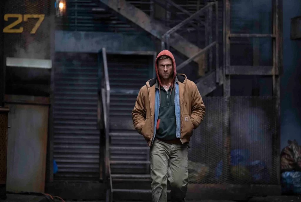 a grungy outdoor location with stairs leading up. A white man wearing a red hood, brown jacket with two shirts underneath, and brown pants is walking toward the screen.