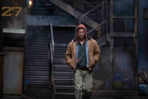 a grungy outdoor location with stairs leading up. A white man wearing a red hood, brown jacket with two shirts underneath, and brown pants is walking toward the screen.
