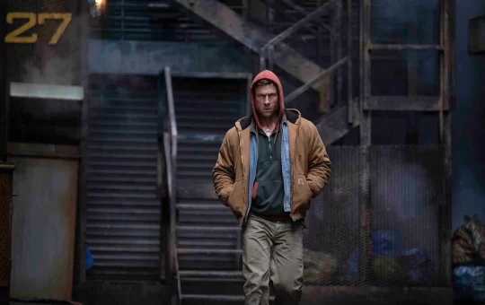 a grungy outdoor location with stairs leading up. A white man wearing a red hood, brown jacket with two shirts underneath, and brown pants is walking toward the screen.