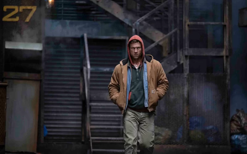 a grungy outdoor location with stairs leading up. A white man wearing a red hood, brown jacket with two shirts underneath, and brown pants is walking toward the screen.