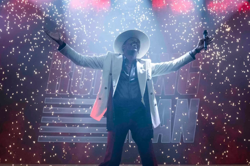a black man wearing a white hat and jacket over a blue suit stands with his arms raised, a microphone in one hand. The background is The Running Man sign and sparks.