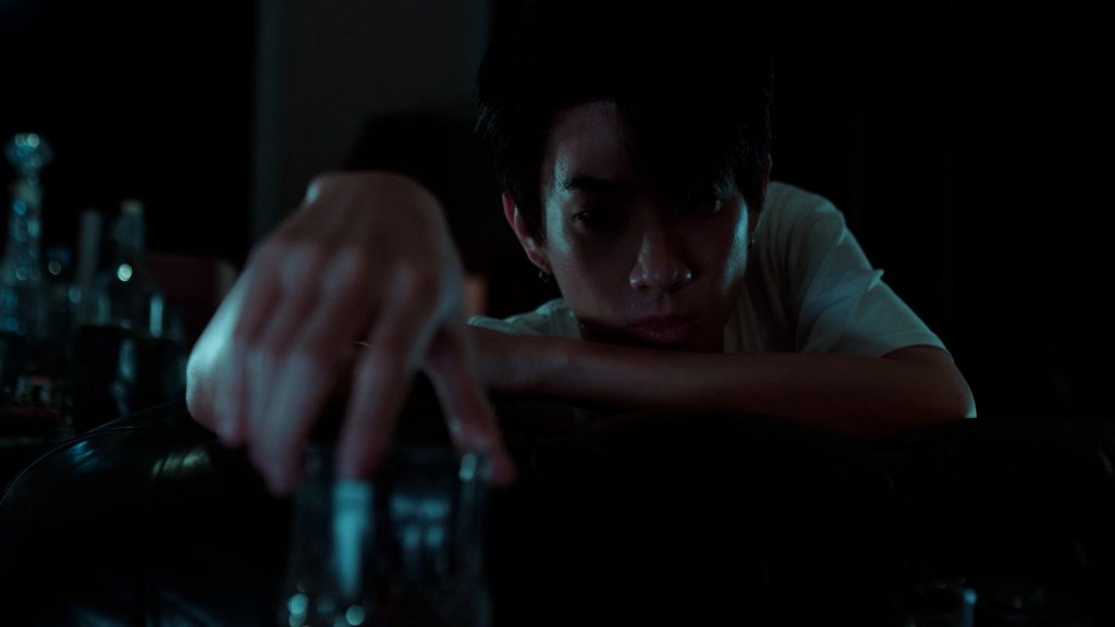 Koh, shadowed in his dark apartment, reaches for the glass of liquor Jira left behind early that day.