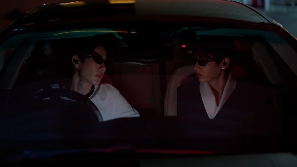 A shot through the windshield of Koh's car as he and Jira sit outside of the awards show. Koh is driving, with Jira in the passenger seat. Despite the fact that it's night, they are both wearing sunglasses.