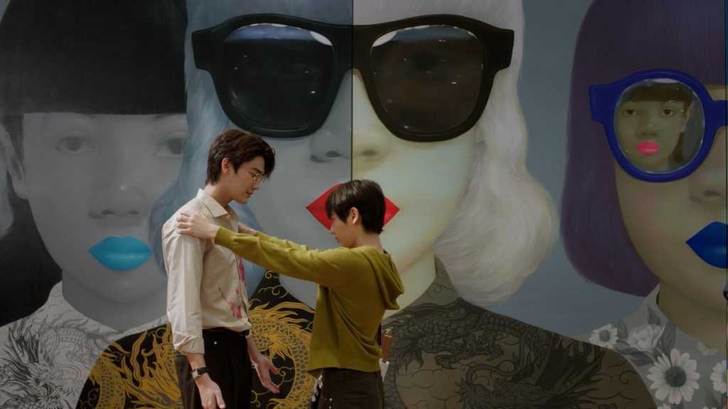 Jira holds Pheem at arm's length, examining the wine-stained/painted shirt he gifted him earlier. They stand in front of a painted mural of women wearing sunglasses.