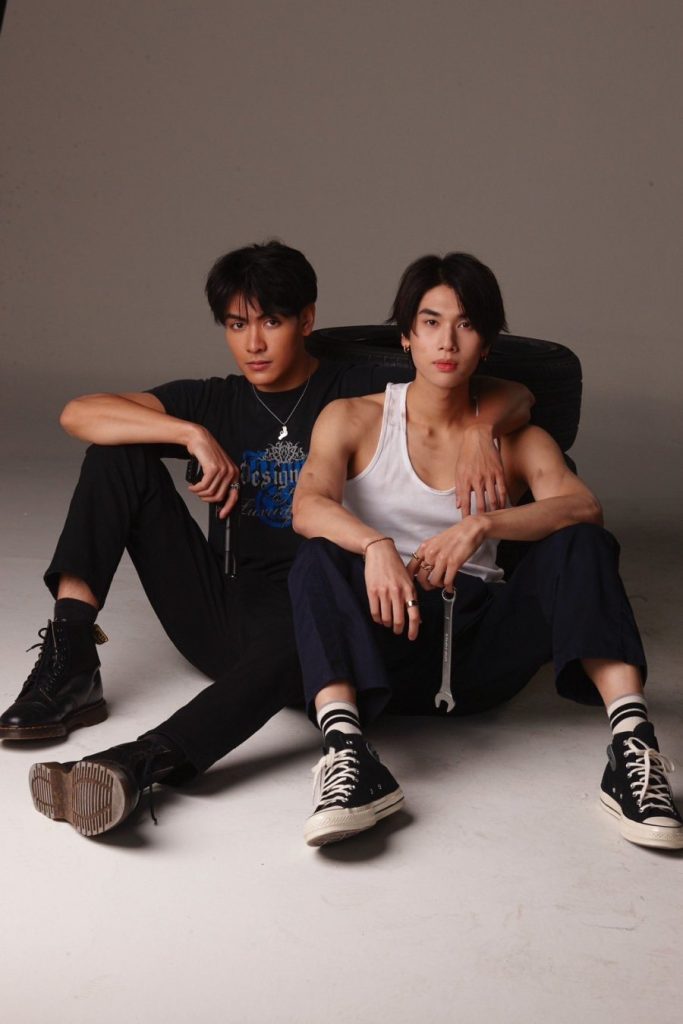 Fadel and Style sit on the floor, leaning against a stack of tires. Style is holding a wrench by dangling it from his pinky. Fadel as one arm around Style and the other hand on his gun.