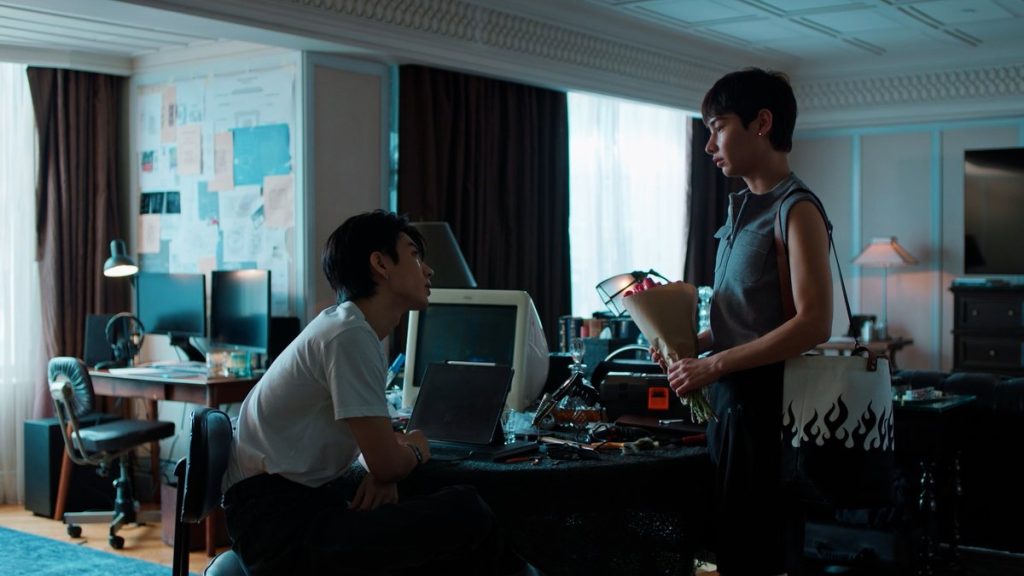 Koh is seated at one of his many desks in his apartment, looking up at Jira. Jira is holding a bouquet of red tulips that Koh bought for him.