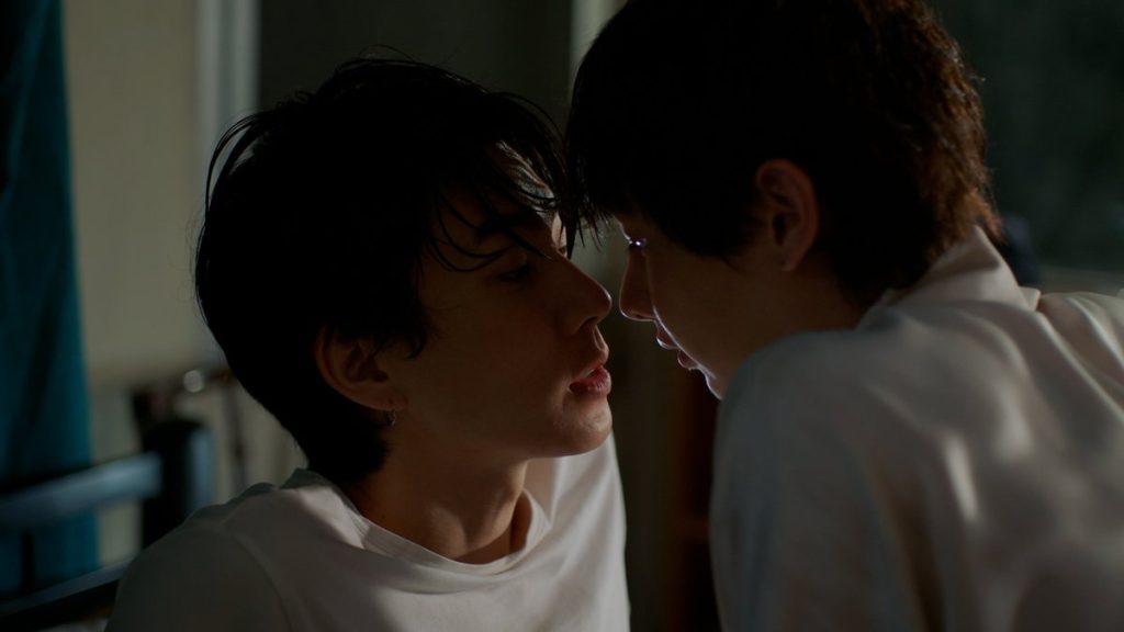 Koh and Jira sit on the bed in Koh's childhood bedroom. Jira is in Koh's lap. They are both leaning in for a kiss.