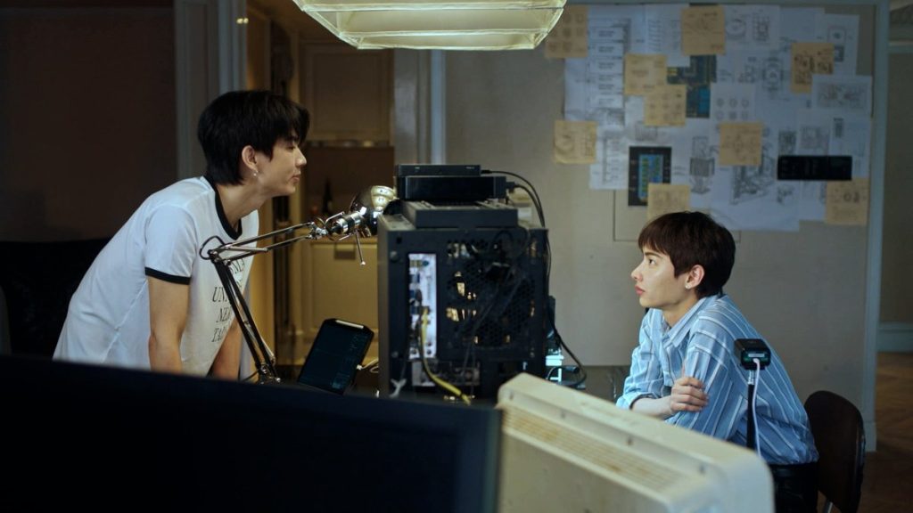 Koh and Jira are across a table from each other. Jira is seated in front of monitors, while Koh is standing opposite, leaning forward over the table.